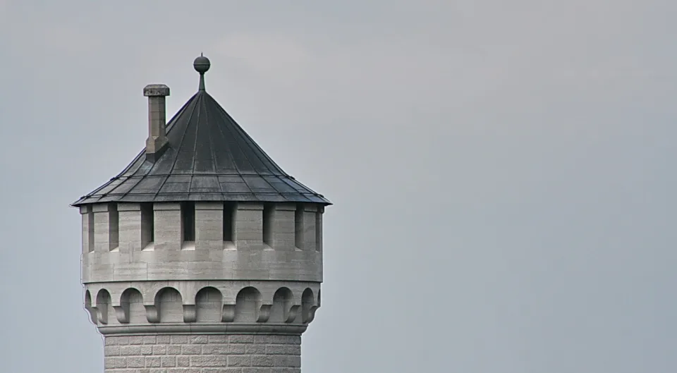 Oberer Teil eines runden Steinturms mit spitzem, dunklem Schieferdach, Zierkugel auf der Spitze und einer Bogengalerie unter dem Dach. Links der Turm, rechts eine leere graue Himmelsfläche.