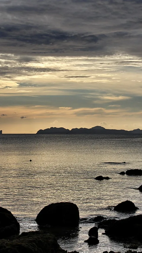 Ruhiges Meer mit dunklen Felsen im Vordergrund, dahinter Inselsilhouetten unter einem Abendhimmel mit Wolken.