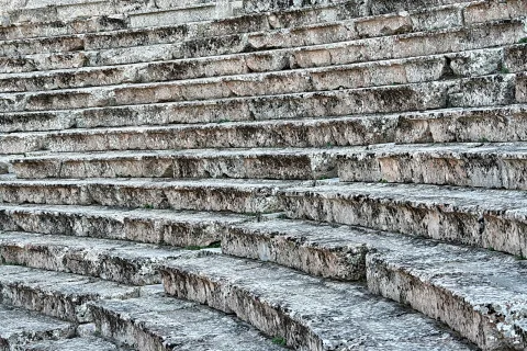 Breite, verwitterte graue Steinstufen eines antiken Theaters, diagonal von unten links nach oben rechts ansteigend.