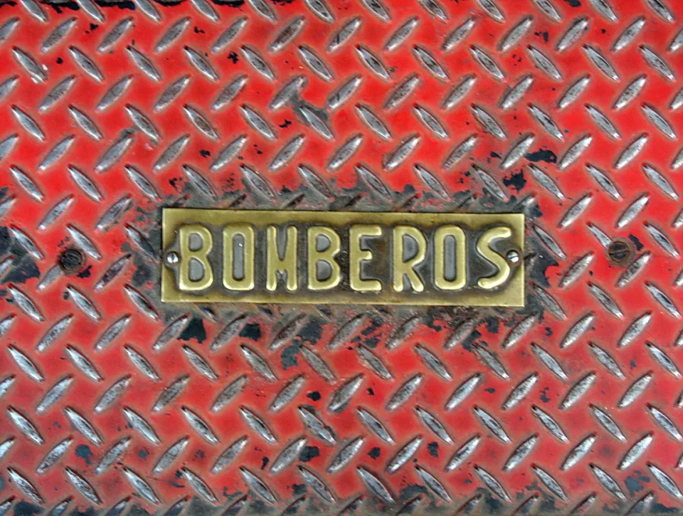 Messingschild mit der Aufschrift BOMBEROS auf einer roten Riffelblech-Oberfläche aus Metall.