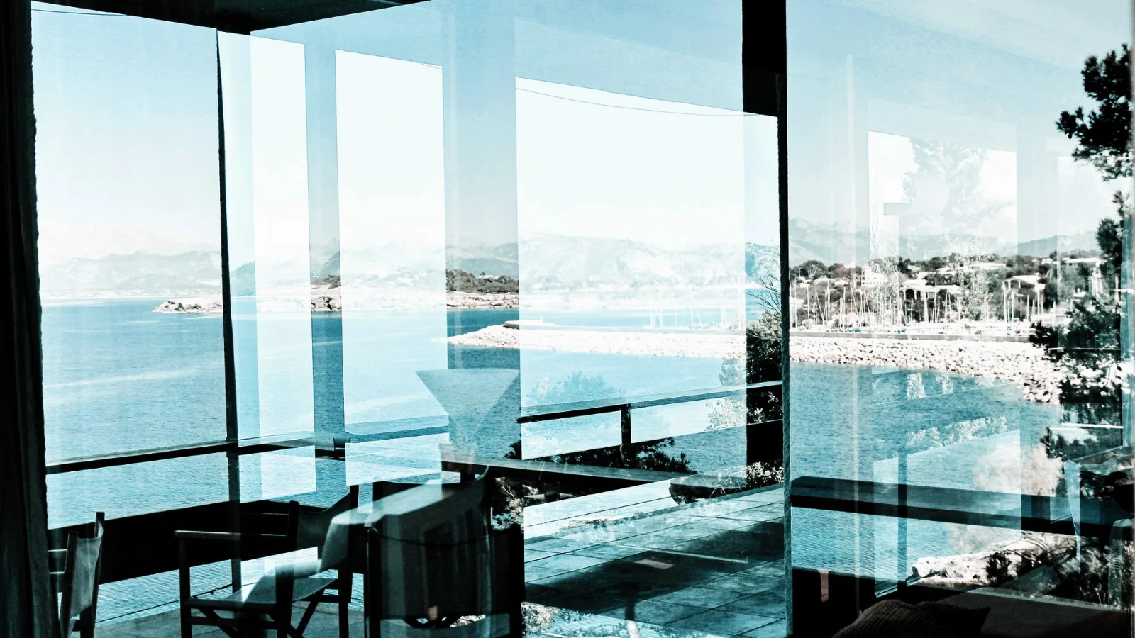 Blick durch große Glasfronten einer modernen Villa auf das Meer, eine Felsküste und einen Hafen mit Booten, im Vordergrund Stühle und Spiegelungen in den Scheiben.