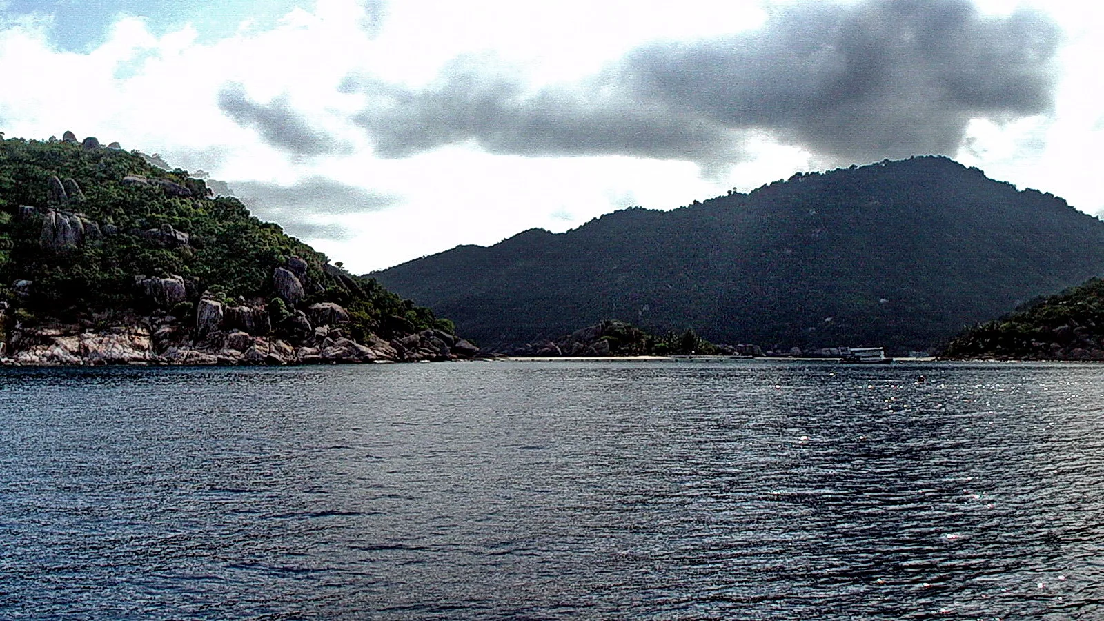 Blick vom Boot aufs ruhige Meer vor bewaldeten Felsinseln und Bergen unter bewölktem Himmel