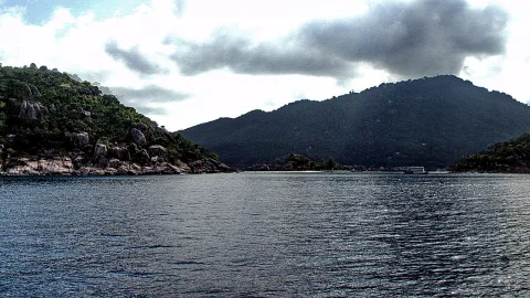 Blick vom Boot aufs ruhige Meer vor bewaldeten Felsinseln und Bergen unter bewölktem Himmel