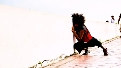 Eine Frau mit Afro-Locken in rotem Top und schwarzer Hose hockt vor einer weißen Mauer und lehnt den Kopf dagegen. Rechts im Hintergrund stehen weitere Menschen auf einem rot gepflasterten Weg.