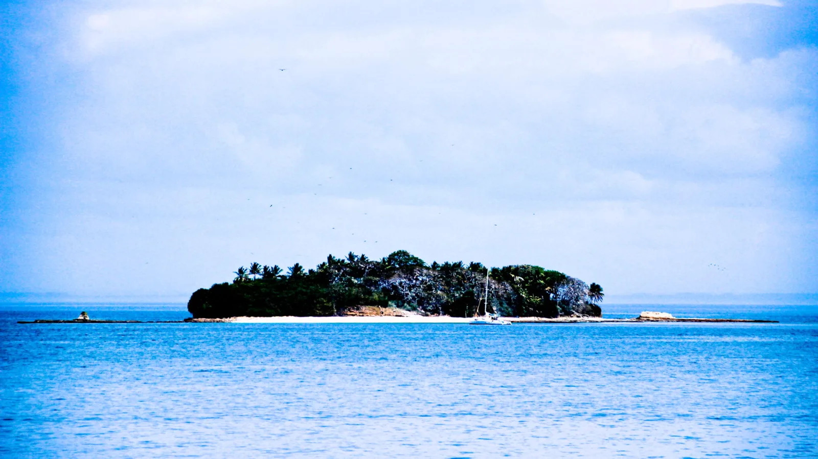 Kleine bewaldete Insel mit Palmen und hellem Sandstrand im blauen Meer, rechts davon ein weißes Segelboot vor Anker, darüber bewölkter Himmel.