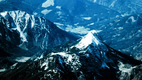 Luftaufnahme einer blau getönten Gebirgslandschaft mit schneebedeckten Gipfeln, darunter ein spitzer Berg im Vordergrund, und einem Tal mit hellen Wiesenflächen