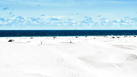 Weite weiße Sanddünen unter blauem Himmel, dahinter das dunkelblaue Meer; im Mittelgrund zwei winzige Personen und einzelne grüne Büsche.