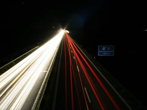 Langzeitbelichtung einer nächtlichen Autobahn: links weiße Scheinwerferspuren, rechts rote Rücklichtspuren, die in die Tiefe zulaufen; rechts ein blaues Hinweisschild mit der Aufschrift Bad Reichenhall 1500 m.