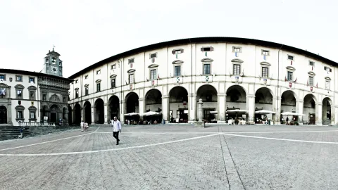 Breiter, gepflasterter Platz vor einer geschwungenen, mehrstöckigen Arkadenfassade mit Rundbögen, links ein Gebäude mit Uhrturm. Ein Mann in weißem Hemd geht über den Platz, unter den Arkaden stehen Sonnenschirme und Tische.