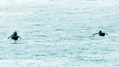 Zwei kleine Ruderboote auf ruhigem, blaugrauem Meer. Links sitzen drei Personen in einem Boot, rechts rudert eine Person allein.