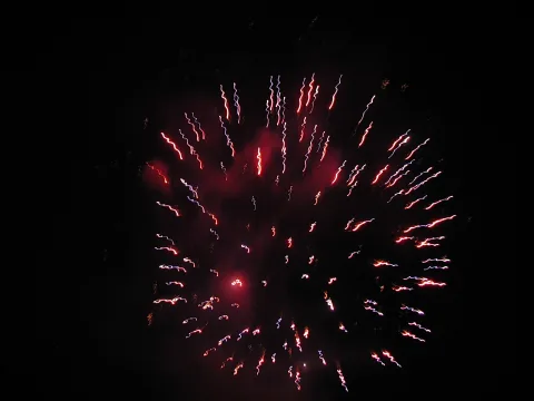 Rote und blaue Lichtspuren von Feuerwerksraketen strahlen vor schwarzem Nachthimmel von einem Mittelpunkt aus.