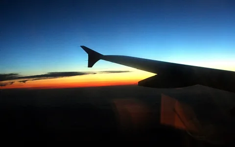 Flugzeugtragfläche vor einem Himmel, der von Dunkelblau über Orange zum Horizont hin verläuft, darunter eine Wolkendecke.