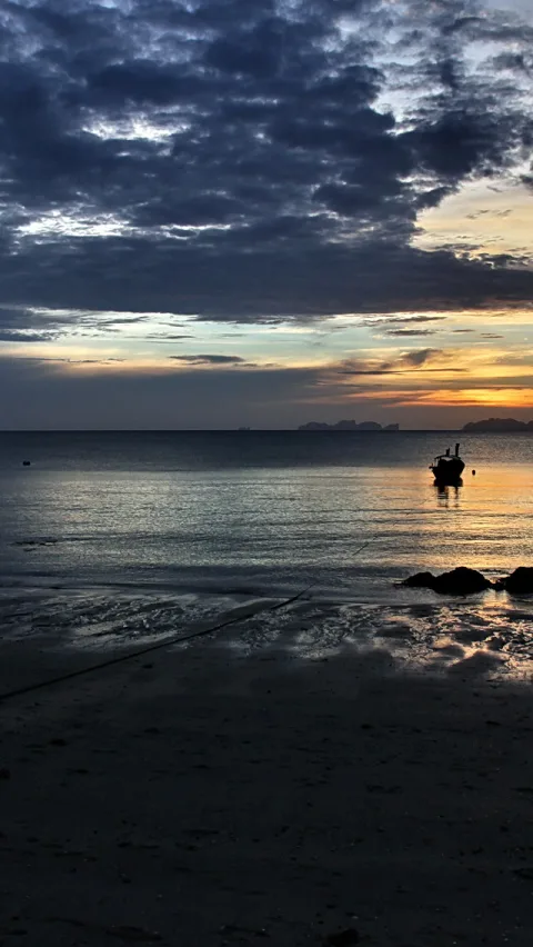 Ein dunkles Boot liegt auf ruhigem Meer vor einem Strand, im Hintergrund ein Sonnenuntergang mit dunklen Wolken und orangefarbenem Horizont.