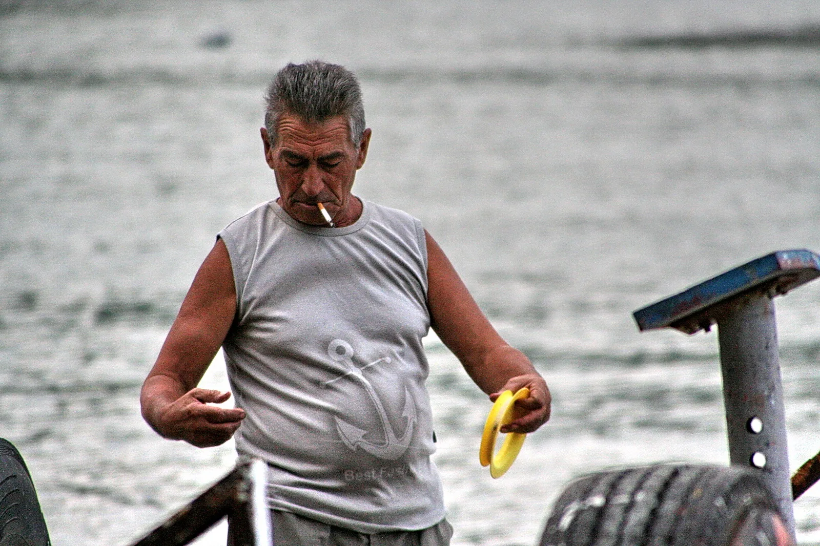 Älterer Mann mit grauen Haaren steht in einem ärmellosen grauen Shirt mit Ankermotiv am Wasser, raucht eine Zigarette und hält einen gelben Ring in der Hand. Im Hintergrund sind Reifen und ein Metallpfosten zu sehen.
