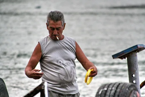 Älterer Mann mit grauen Haaren steht in einem ärmellosen grauen Shirt mit Ankermotiv am Wasser, raucht eine Zigarette und hält einen gelben Ring in der Hand. Im Hintergrund sind Reifen und ein Metallpfosten zu sehen.