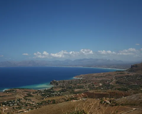 Weiter Blick von einer Anhöhe über eine tiefblaue Bucht mit türkisfarbenem Wasser, Küstenort mit Häusern und Feldern, dahinter Bergketten unter blauem Himmel mit wenigen weißen Wolken.