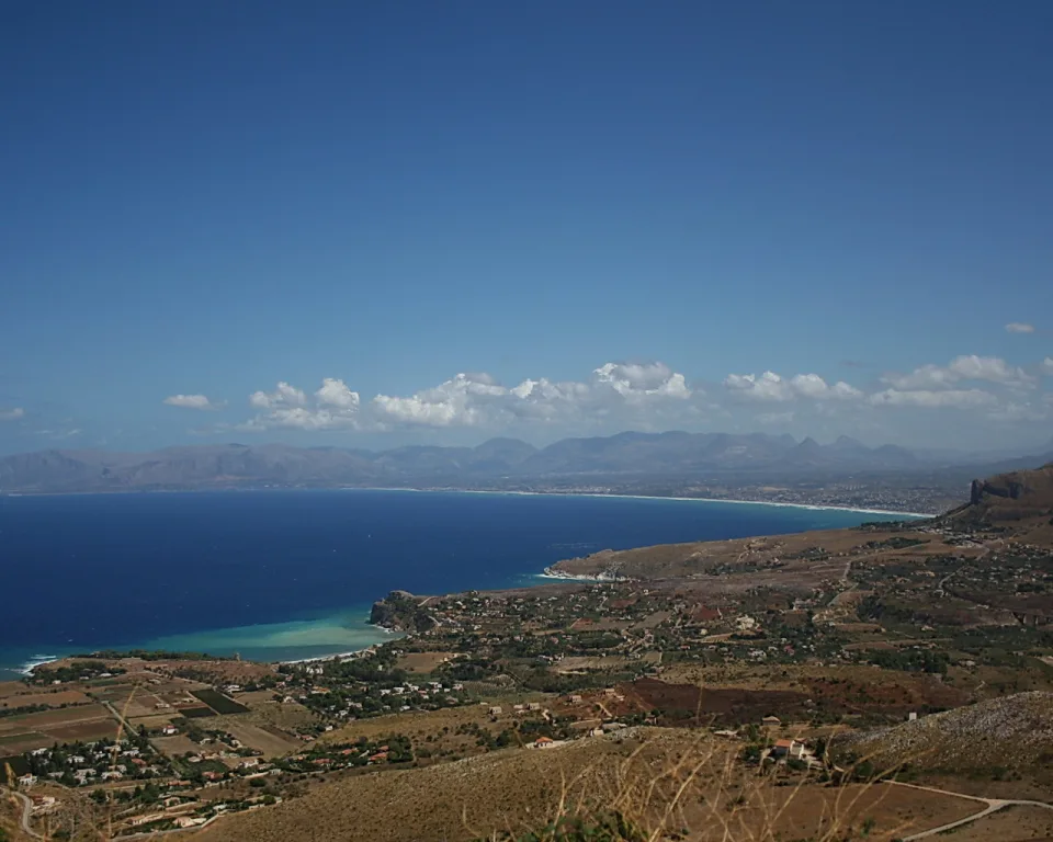 Weiter Blick von einer Anhöhe über eine tiefblaue Bucht mit türkisfarbenem Wasser, Küstenort mit Häusern und Feldern, dahinter Bergketten unter blauem Himmel mit wenigen weißen Wolken.