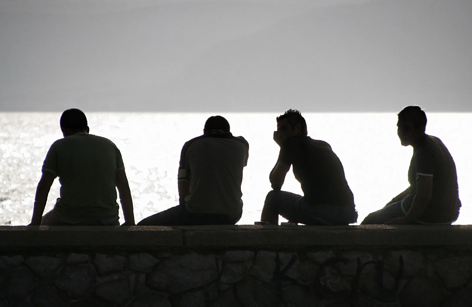 Vier Männer sitzen nebeneinander auf einer Steinmauer am Meer, als dunkle Silhouetten vor hellem Gegenlicht.