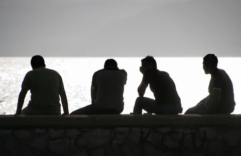 Vier Männer sitzen nebeneinander auf einer Steinmauer am Meer, als dunkle Silhouetten vor hellem Gegenlicht.