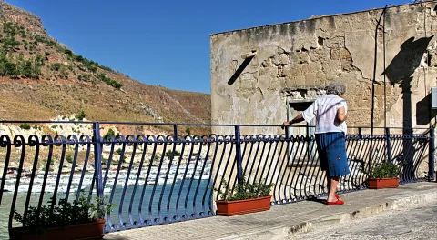 Eine ältere Frau mit weißer Bluse und blauem Rock lehnt am blauen schmiedeeisernen Geländer und blickt auf einen Hafen mit Booten. Links steigt ein karger Hügel an, rechts steht eine verwitterte Steinmauer.