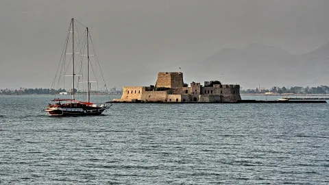 Kleine steinerne Festung mitten im Meer, links davon ein Segelboot mit roten Akzenten, im Hintergrund dunkle Berge unter bewölktem Himmel.