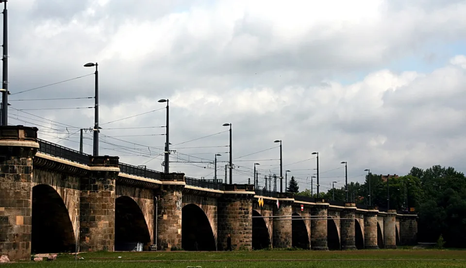 Lange Steinbogenbrücke mit Laternen und Oberleitungen unter bewölktem Himmel, im Vordergrund eine grüne Wiese.