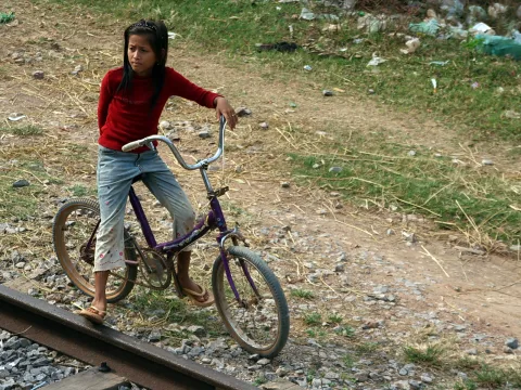 Ein Mädchen mit rotem Langarmshirt und Jeans steht auf einem lila Fahrrad neben einem Bahngleis, im Hintergrund eine Böschung mit Müll und Gras.