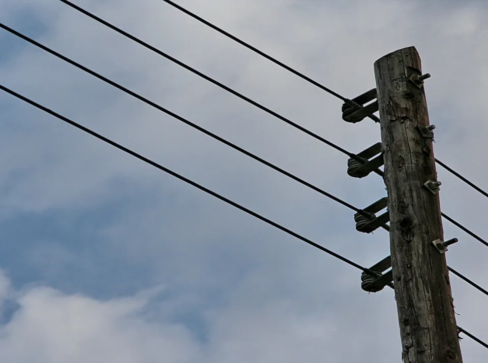 Holzmast einer Stromleitung von unten fotografiert, rechts im Bild, mit vier schräg verlaufenden Drähten vor einem grau-blauen Wolkenhimmel.