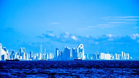 Skyline einer Großstadt mit dichten Hochhäusern am Meer unter blauem Himmel, im Vordergrund dunkelblaues, leicht bewegtes Wasser und zwei Schiffe.