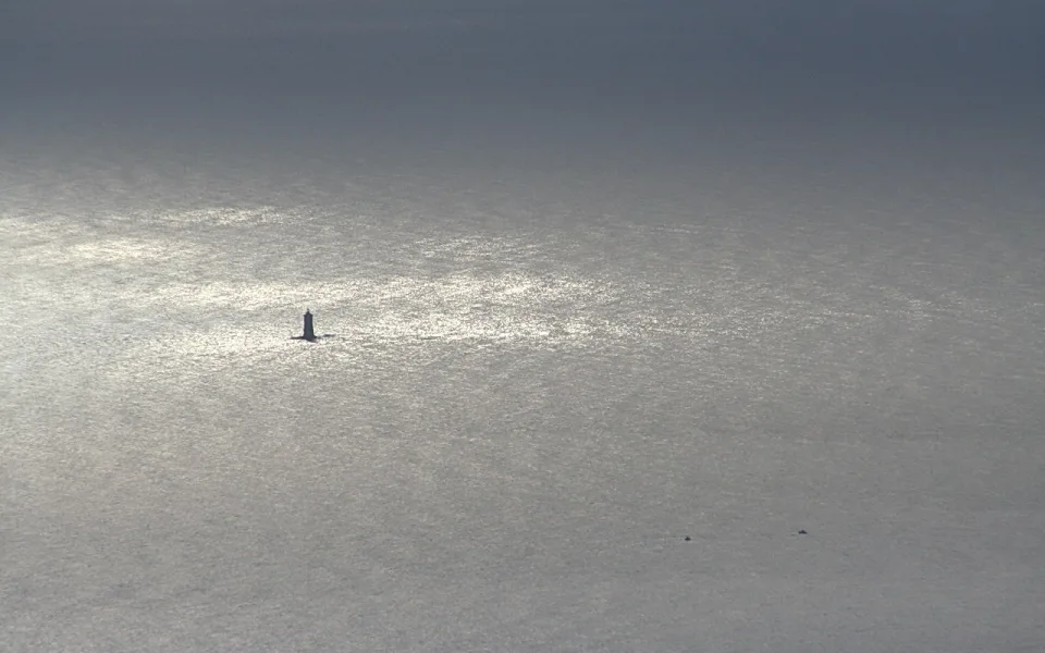 Ein kleiner Leuchtturm steht allein auf einer glänzenden, grauen Meeresfläche, auf der sich helles Licht spiegelt.