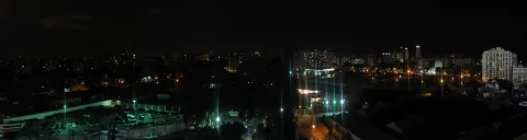 Panoramablick auf eine nächtliche Großstadt mit beleuchteten Straßen, einem Parkplatz mit geparkten Autos im Vordergrund und hell erleuchteten Hochhäusern am rechten Bildrand unter dunklem Himmel.