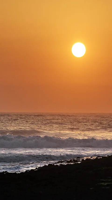 Orangefarbener Sonnenuntergang über dem Meer, die Sonne steht knapp über dem Horizont, im Vordergrund brechen Wellen auf dunklen Felsen.