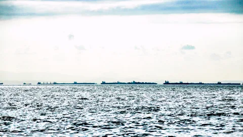 Mehrere große Frachtschiffe und Tanker liegen als dunkle Silhouetten am Horizont. Im Vordergrund wellt sich die graublaue Meeresoberfläche unter einem hellen, bewölkten Himmel.