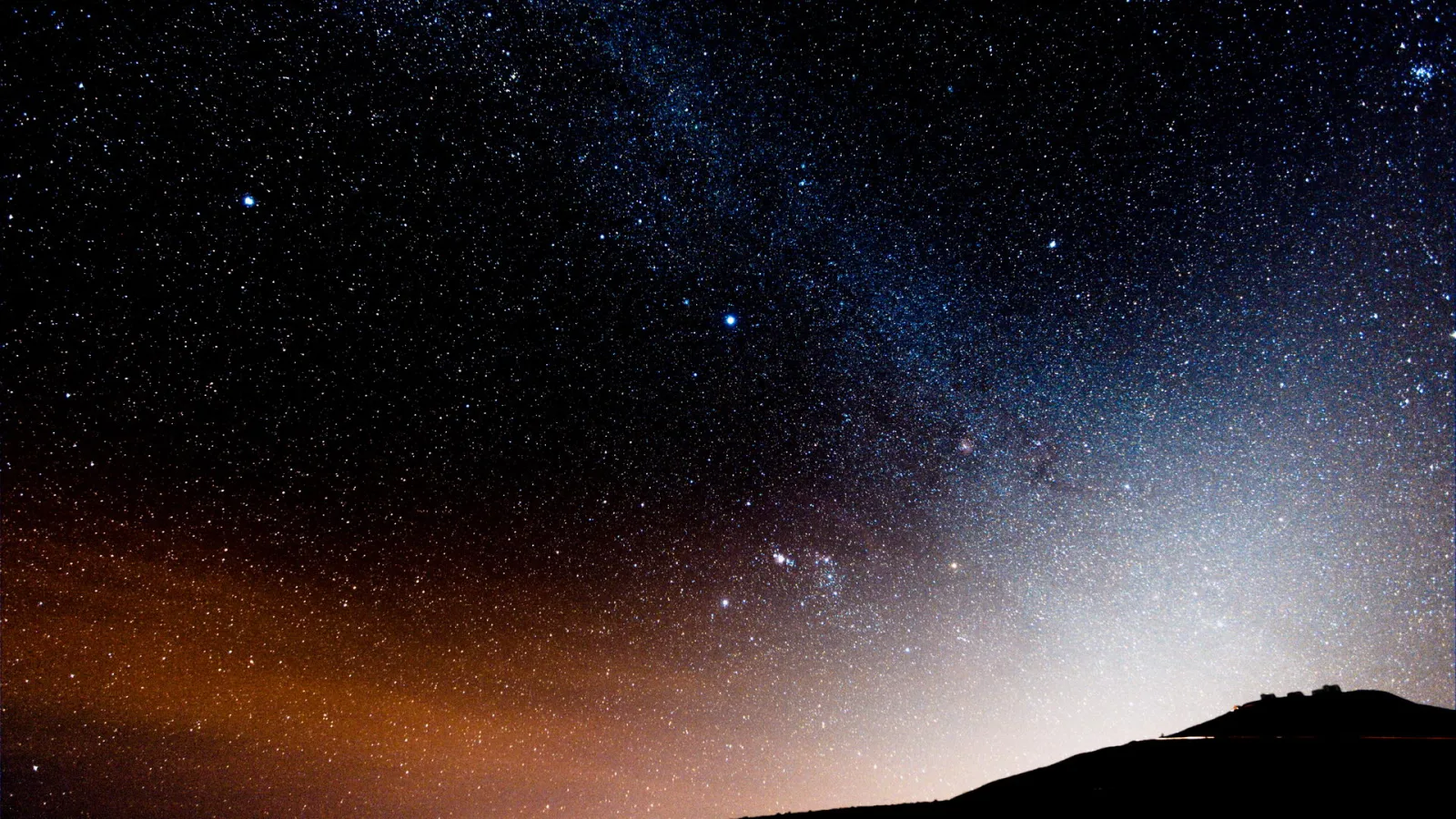 Nachthimmel mit dichtem Sternenfeld und einem hellen Band der Milchstraße. Unten rechts zeichnet sich eine dunkle Hügelkuppe mit einem kleinen Gebäude ab, links am Horizont ein orangefarbener Lichtschimmer.