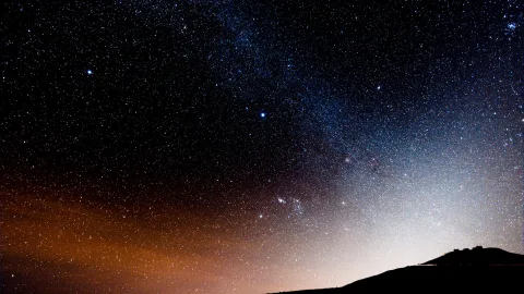 Nachthimmel mit dichtem Sternenfeld und einem hellen Band der Milchstraße. Unten rechts zeichnet sich eine dunkle Hügelkuppe mit einem kleinen Gebäude ab, links am Horizont ein orangefarbener Lichtschimmer.