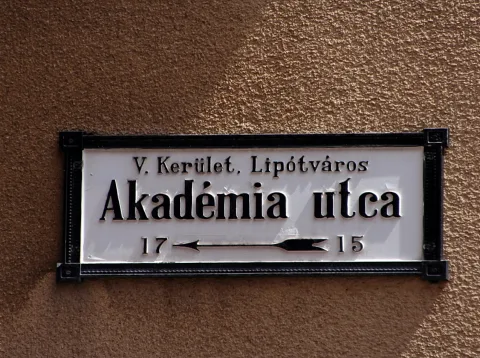 Weißes Straßenschild mit schwarzer Schrift und schwarzem Rahmen an einer braunen Putzwand, zeigt Akadémia utca, V. Kerület, Lipótváros sowie die Hausnummern 17 und 15 mit Pfeil.