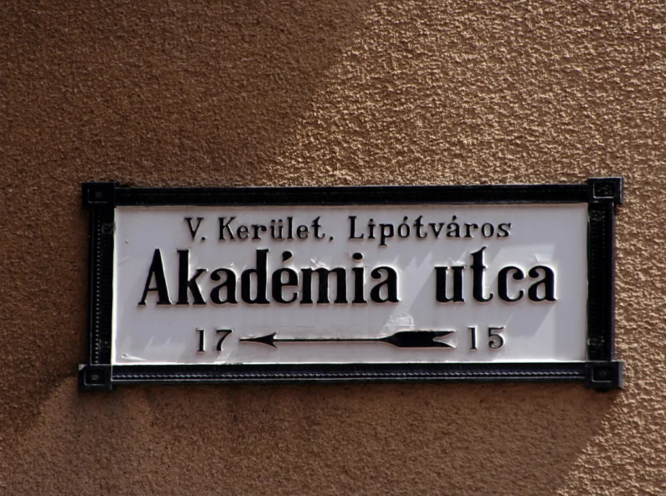 Weißes Straßenschild mit schwarzer Schrift und schwarzem Rahmen an einer braunen Putzwand, zeigt Akadémia utca, V. Kerület, Lipótváros sowie die Hausnummern 17 und 15 mit Pfeil.