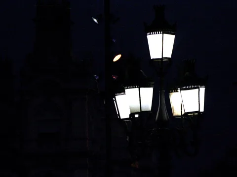 Mehrarmige schmiedeeiserne Straßenlaternen leuchten in der Nacht vor einer dunklen Silhouette eines Gebäudes mit Turm.