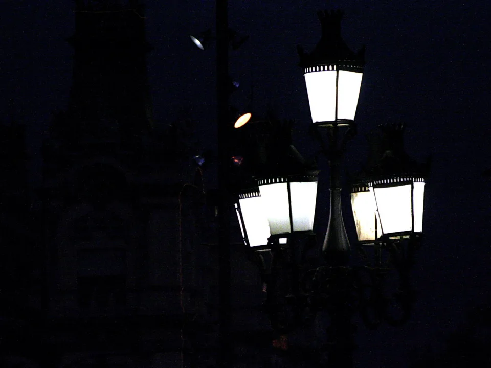 Mehrarmige schmiedeeiserne Straßenlaternen leuchten in der Nacht vor einer dunklen Silhouette eines Gebäudes mit Turm.