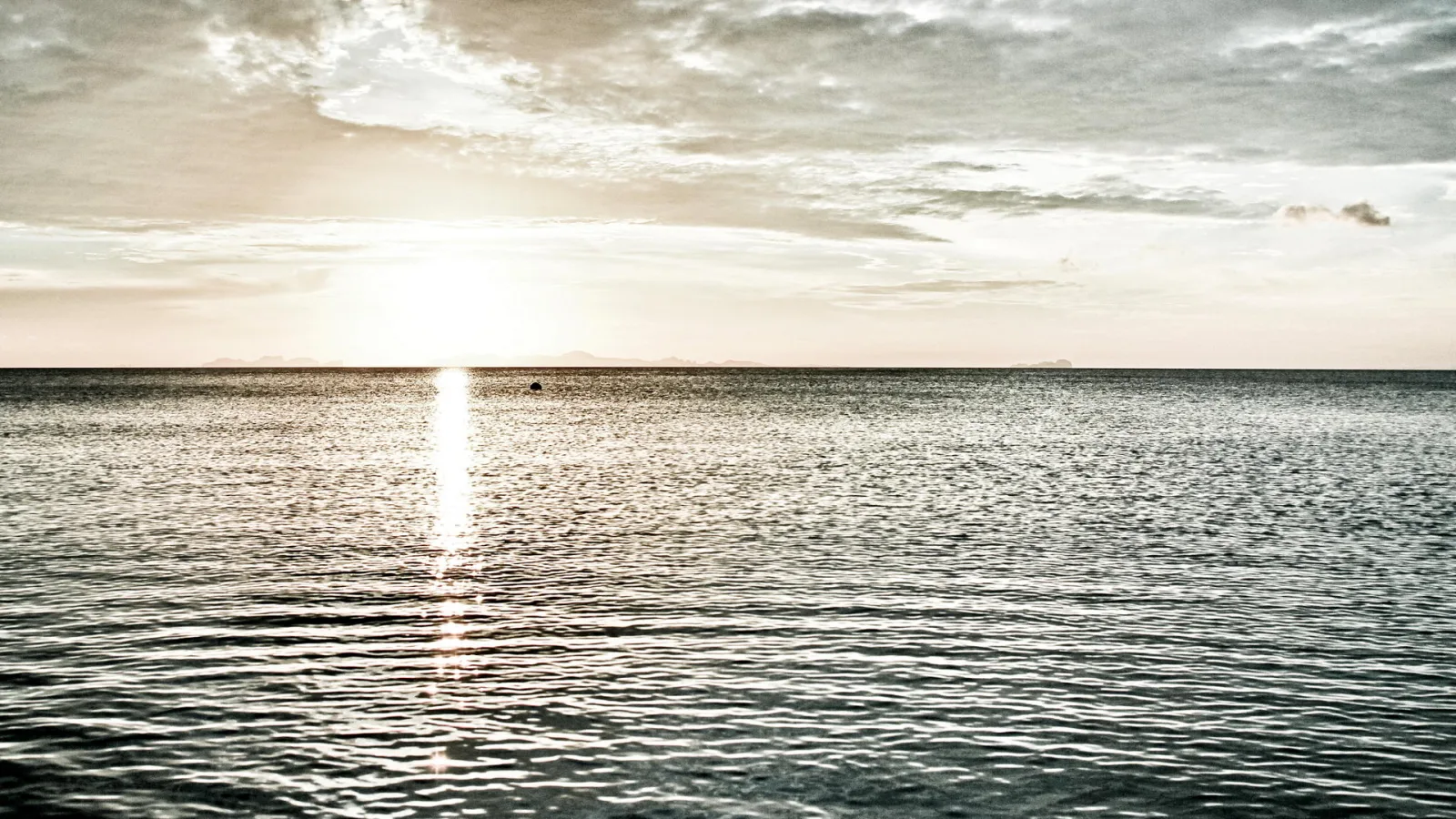 Ruhige Meeresoberfläche bei Sonnenuntergang, die Sonne steht tief am Horizont und spiegelt sich als helle Bahn auf dem Wasser, darüber ein bewölkter Himmel.