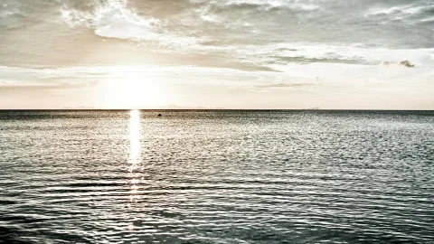Ruhige Meeresoberfläche bei Sonnenuntergang, die Sonne steht tief am Horizont und spiegelt sich als helle Bahn auf dem Wasser, darüber ein bewölkter Himmel.