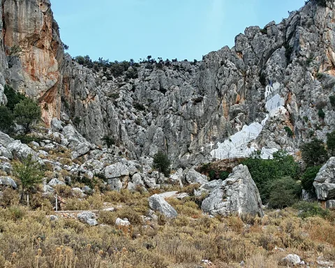 Steile Felswand mit einer weiß gefassten Treppe und kleinen Kreuzen, davor Geröll und trockenes Gras unter blauem Himmel.