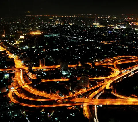 Luftaufnahme einer nächtlichen Stadt mit mehrstöckigen Autobahnkreuzungen, deren Fahrspuren als goldene Lichtbänder zu sehen sind, dahinter ein dichtes Lichtermeer von Hochhäusern.