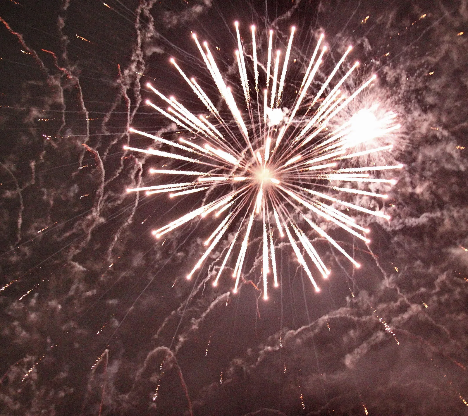 Ein weißes Feuerwerk mit radial abstrahlenden Funkenbahnen vor dunklem Nachthimmel, dazwischen Rauchschwaden und Glutpunkte.