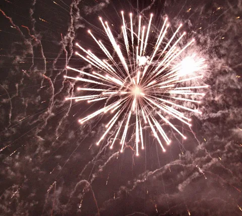 Ein weißes Feuerwerk mit radial abstrahlenden Funkenbahnen vor dunklem Nachthimmel, dazwischen Rauchschwaden und Glutpunkte.