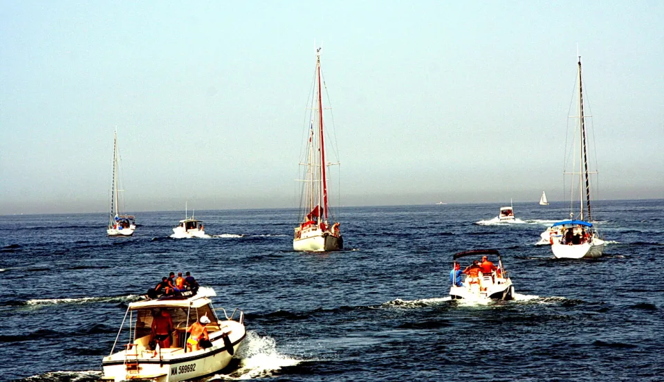 Mehrere Segel- und Motorboote fahren bei Sonnenschein über ein blaues Meer, im Vordergrund ein Motorboot mit Personen in Schwimmwesten, dahinter zwei Segelboote mit hohen Masten.