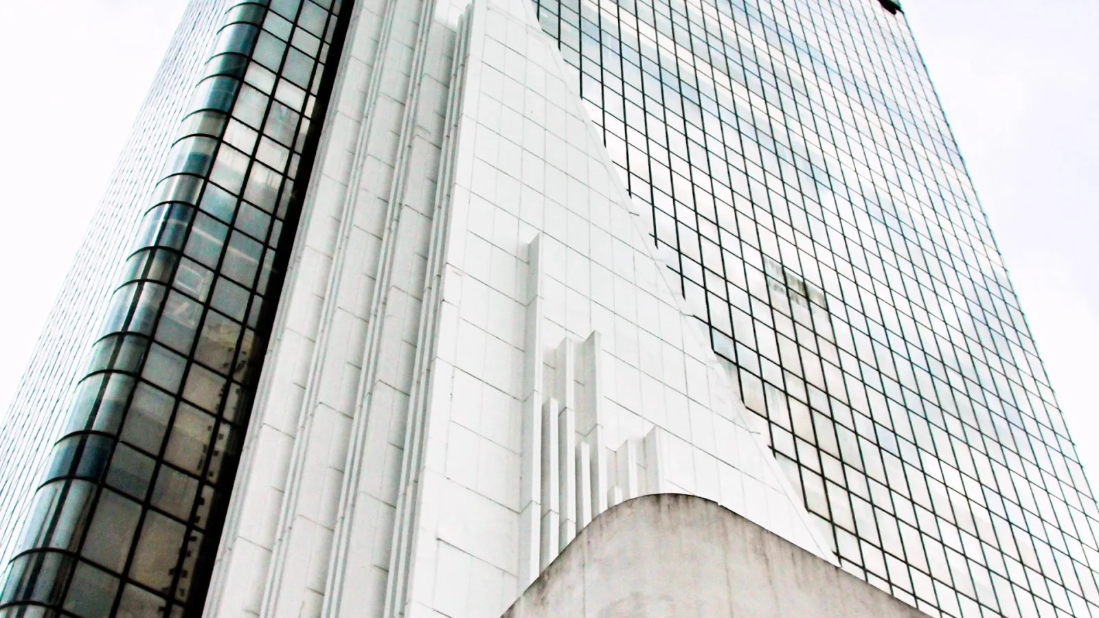 Blick von unten auf ein Hochhaus mit weißer Kachelfassade und einer Glasfront mit feinem Gittermuster vor hellem Himmel.