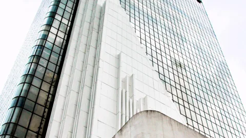 Blick von unten auf ein Hochhaus mit weißer Kachelfassade und einer Glasfront mit feinem Gittermuster vor hellem Himmel.
