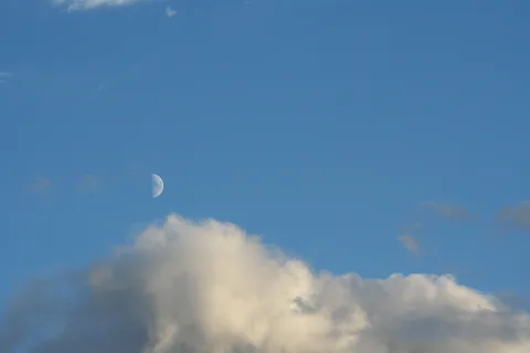 Ein halber Mond steht am blauen Himmel über einer großen, hellen Wolkenbank, die den unteren Bildrand einnimmt.