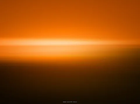 Verschwommene Aufnahme mit leuchtend orangefarbenem Himmel, einem hellen horizontalen Lichtstreifen in der Mitte und einer dunklen, braunen Fläche im unteren Bildbereich.